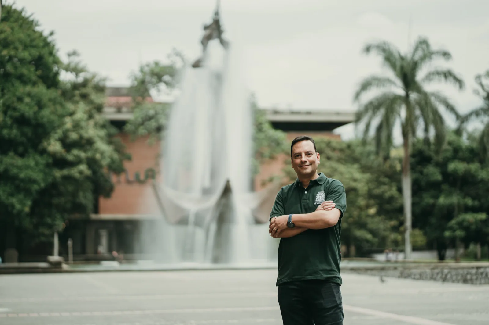 Brazos cruzados frente a la fuente del campus, postura segura.
